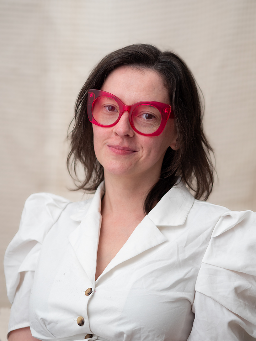 Jessica Leonard, in large pink-framed glasses and a white shirt, half smiling at the camera.