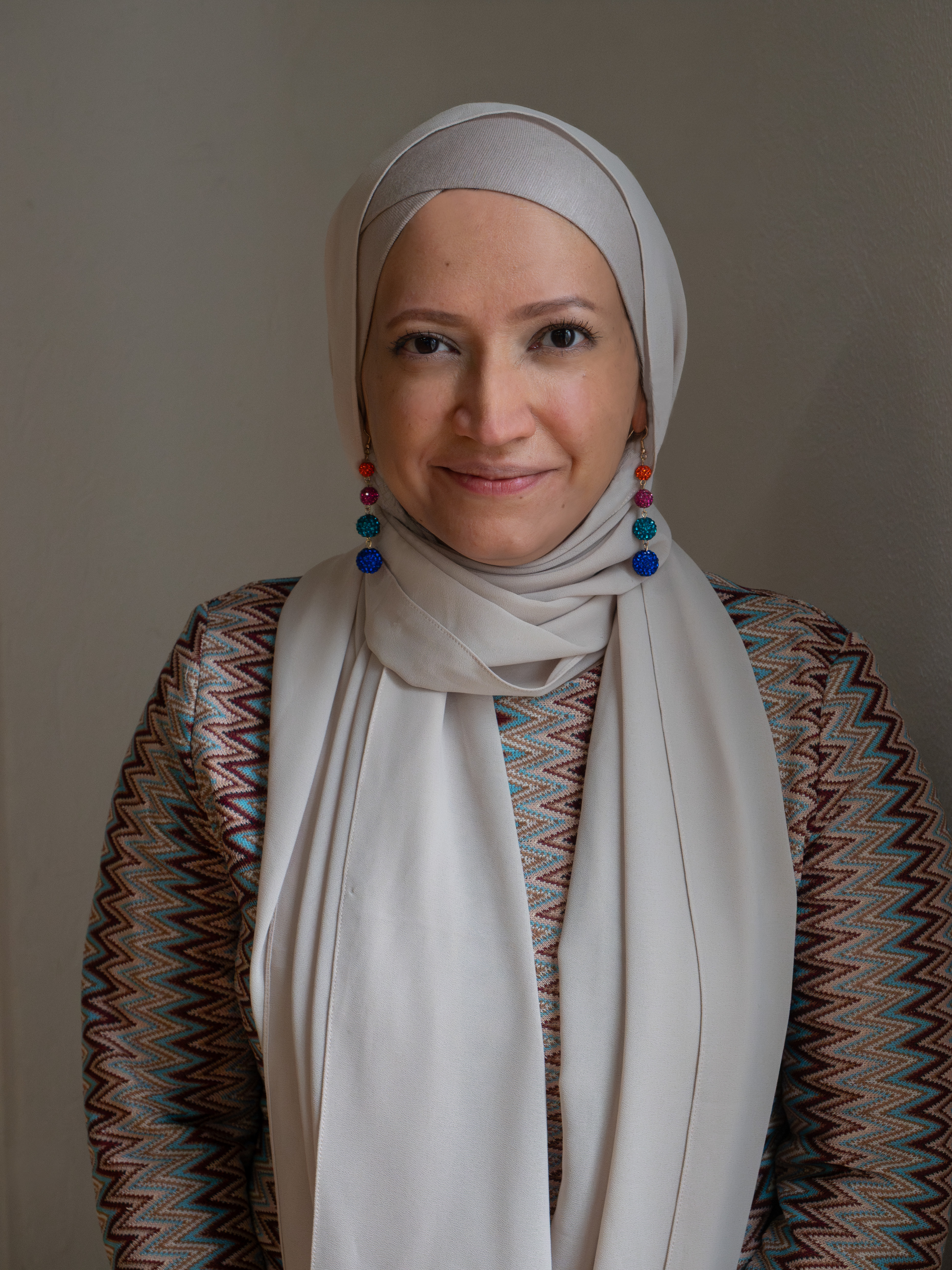 Shazma Gaffoor, in a cream hijab and a zigzag-patterned jacket with bright beaded drop earrings, smiling at the camera.